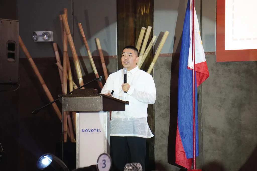 A speaker in a traditional Barong Tagalog addressing the audience from the Novotel podium during the GGDI event.