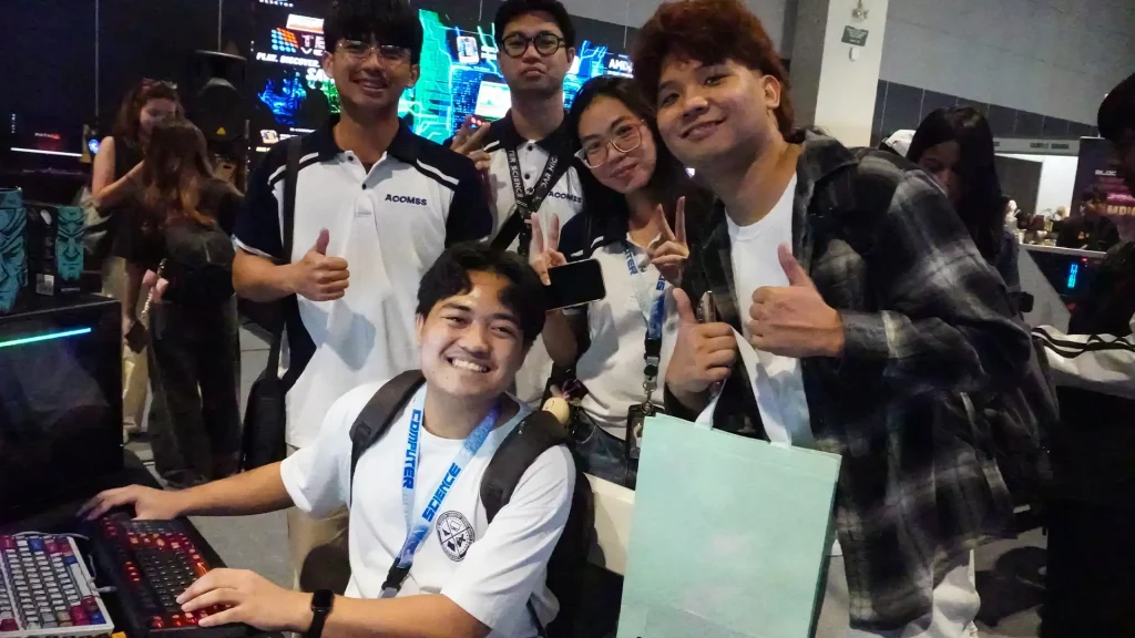 A group of smiling tech convention attendees and computer science students posing together at a mechanical keyboard and gaming PC booth setup.