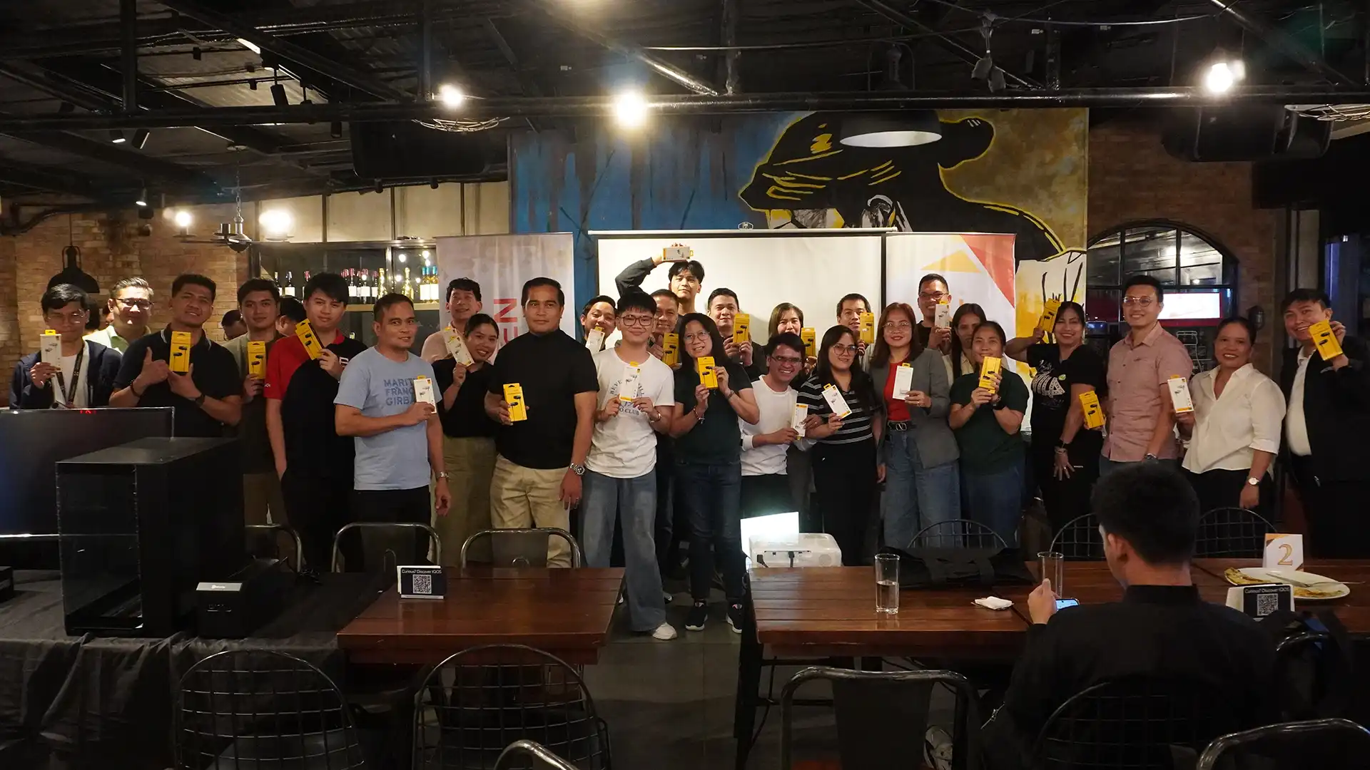 A large group of event attendees standing together and holding up yellow product retail boxes for a group photo.