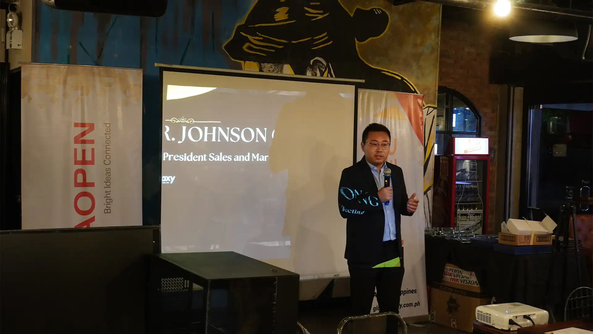 Corporate executive speaking into a microphone on stage next to a presentation screen and an AOPEN banner.
