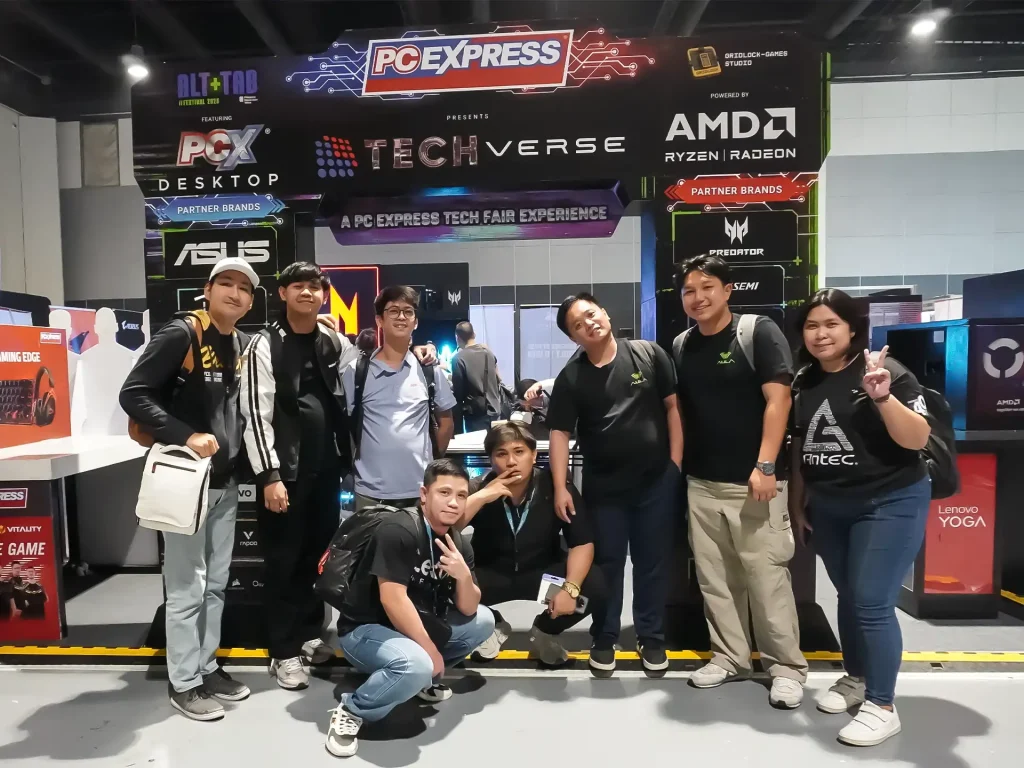 A group of eight event organizers and attendees posing together under the main illuminated entrance archway of the PC Express Tech Verse experience.